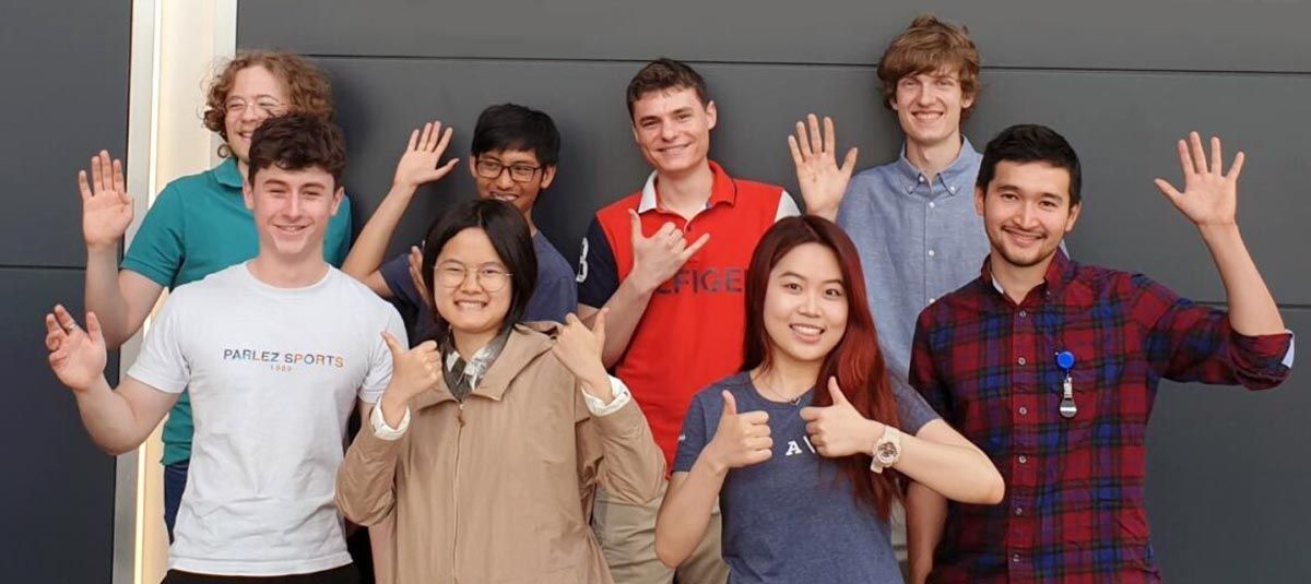 Eight MathWorks interns stand in two rows of four in the Cambridge, UK, office and smile and wave at the camera.