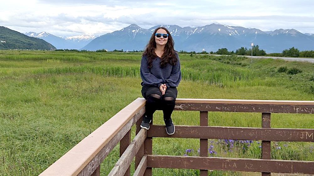 Young woman wearing sunglasses sitting on a fence with grassy area and mountains in the background