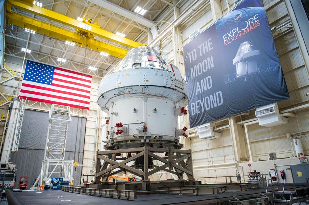 La nave espacial Orion dentro de una instalación de gran tamaño, con la bandera de los Estados Unidos en una pared y una pancarta en otra en la que se lee “A la Luna y más allá”.